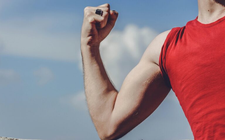 Close-up of a flexed arm in a red shirt against a clear blue sky, symbolizing strength and empowerment.