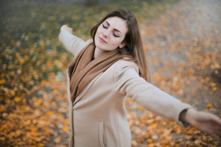 A woman with eyes closed embraces the autumn season amidst fallen leaves.